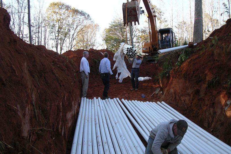 Crew laying pipe in a deep trench with an excavator working behind them